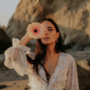 A stylish woman in a floral dress holds a pink flower near her face on a serene beach at sunset.